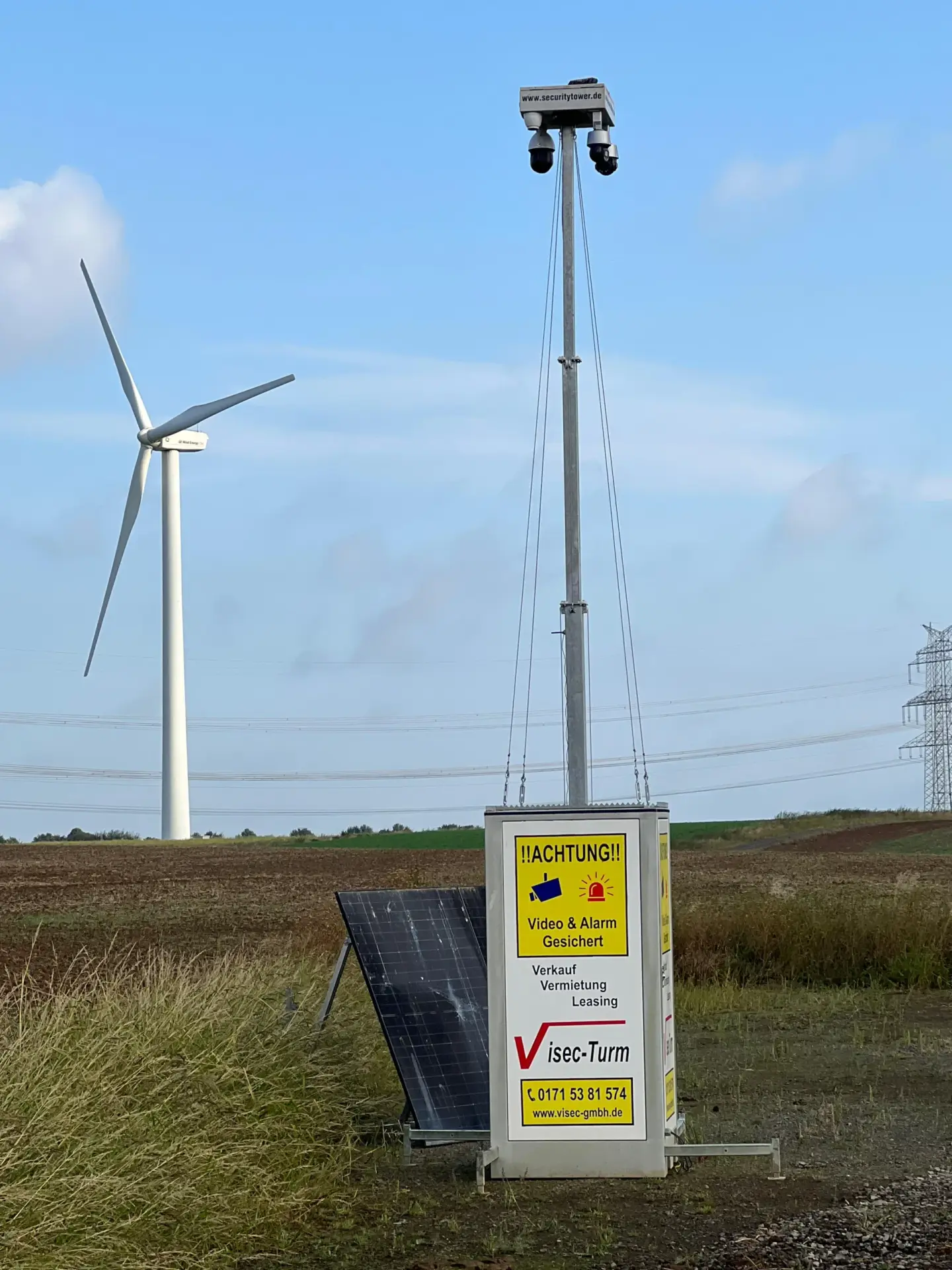 Mobiler Sicherheits-Turm mit Solarzellen und Kameras für Videoüberwachung, aufgestellt in ländlicher Umgebung neben Windkraftanlage.