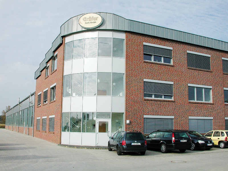Modernes Bürogebäude aus roten Ziegeln mit großen Glasfenstern, umgeben von geparkten Autos.