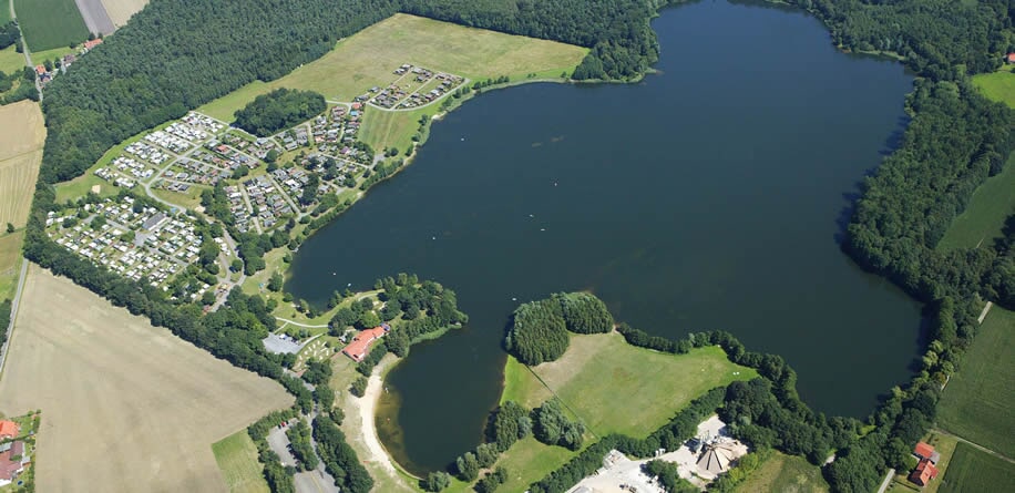Luftaufnahme eines großen Sees, umgeben von Wäldern, einer Siedlung und landwirtschaftlichen Flächen.
