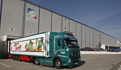 Ein grüner LKW mit "Fresh Harvest"-Auflieger steht vor einem modernen Lagerhaus mit "Godeland"-Logo.