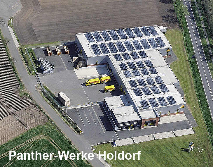 Luftaufnahme eines Industriegebäudes mit Solarpaneelen, gelben Lastwagen und gestapelten Materialien.