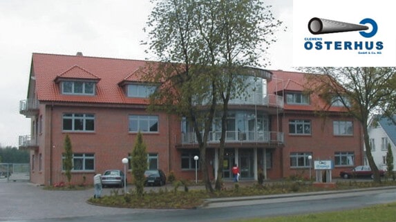 Modernes Bürogebäude aus rotem Backstein mit Ziegeldach, Glasfront und Parkplatz, bewölkter Himmel.