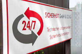 Schild eines Sicherheitsunternehmens mit roten und grauen Elementen, das 24/7-Dienstleistungen anbietet.