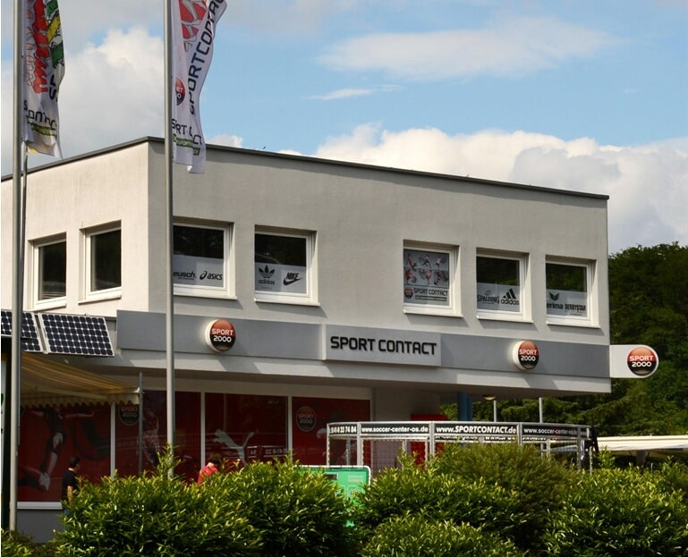 Ein modernes Sportgeschäft mit großen Fenstern, Markenlogos und Fahnen in grüner Umgebung.
