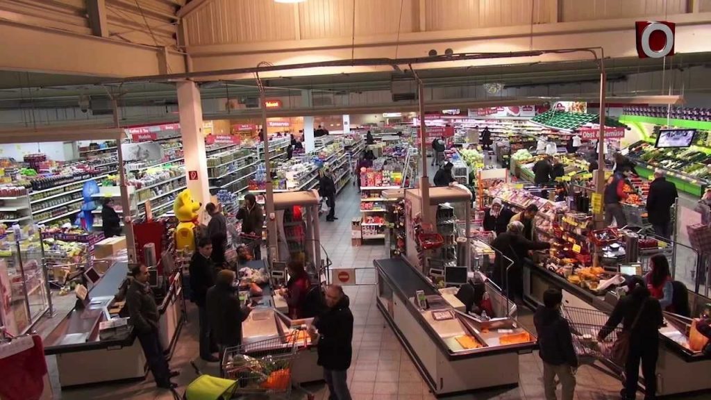 Ein geschäftiger Supermarkt mit Kunden an den Kassen und vollen Regalen mit bunten Produkten.
