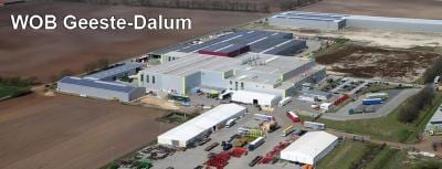 Luftaufnahme eines Industriegebiets mit großen Hallen, Fahrzeugen und Containern, umgeben von Feldern.