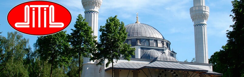 Eine weiße Moschee mit zwei Minaretten, umgeben von grünen Bäumen, unter einem klaren blauen Himmel.
