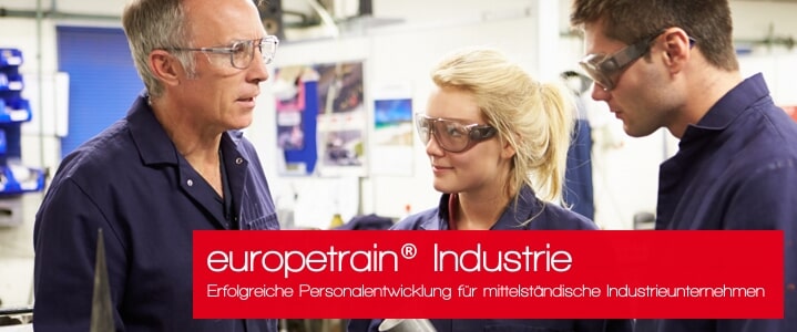 Drei Personen in Arbeitskleidung und Schutzbrillen diskutieren in einer industriellen Umgebung.